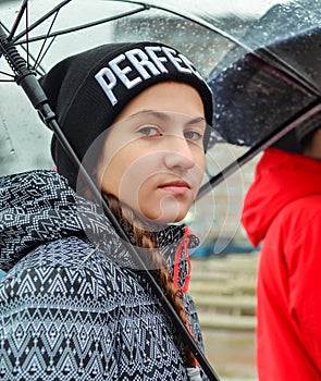 Lifestyle portrait of teenager girl under transparant umbrella wearing black knitted hat and casual rain jacket at rainy