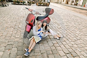 Lifestyle portrait of two Caucasian women friends in the city, using smartphone while sitting on the pavement with