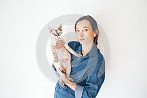 Lifestyle portrait of a woman in blue jeans shirt with wondering expression is holding his curious Devon Rex cat. Curious kitty in