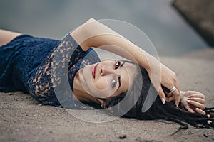 Lifestyle portrait of a woman brunette on background of the lake lying in sand on a cloudy day. Romantic, gentle, mystical