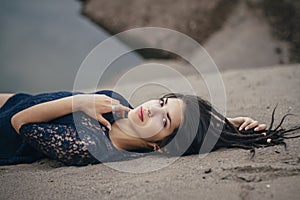 Lifestyle portrait of a woman brunette on background of the lake lying in sand on a cloudy day. Romantic, gentle, mystical