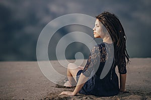 Lifestyle portrait of a woman brunettes in background of lake sitting in sand on a cloudy day. Romantic, gentle, mystical