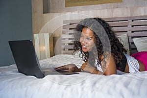 Lifestyle portrait of young beautiful and happy black hispanic woman at home bedroom lying cheerful on bed networking using laptop