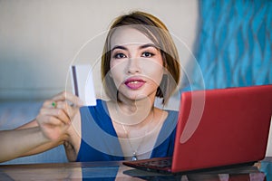 Lifestyle portrait of young beautiful and happy girl at home living room holding credit card using laptop computer for banking an