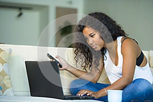 Lifestyle portrait of young happy and beautiful black afro American woman using internet mobile phone while working on laptop com