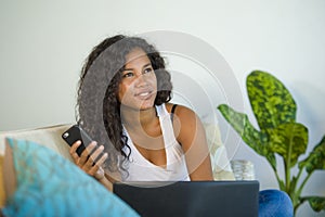 Lifestyle portrait of young happy and beautiful black afro American woman using internet mobile phone while working on laptop comp