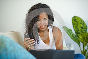 Lifestyle portrait of young happy and beautiful black latin American woman using internet mobile phone while working on laptop co