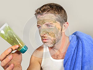 Lifestyle portrait of young messy funny man in bathroom with green face looking at cream male beauty product applying facial mask