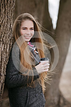 Lifestyle portrait of young and pretty adult woman with gorgeous long hair posing near tree trunk with coffee cup in city park wit