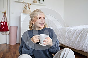 Lifestyle portrait of young woman sitting on bedroom floor with cup of tea, drinking from big white mug and looking