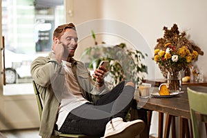 Lifestyle shot of handsome young man, looking at his mobile phone, listening to music in wireless earphones, sitting in