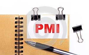 On a light background, a brown notebook with a pen, black clips and a white card with the text PMI Project Management Institute