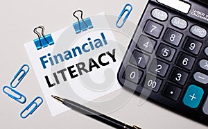 On a light background, a calculator, a pen, blue paper clips and a sheet of paper with the text FINANCIAL LITERACY. View from photo