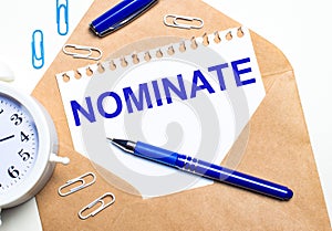 On a light background, a craft envelope, an alarm clock, paper clips, a blue pen and a sheet of paper with the text NOMINATE