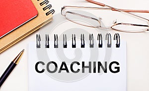 On a light background - glasses in gold frames, a pen, brown and red notepads and a white notebook with the text COACHING.