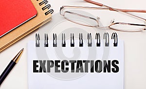 On a light background - glasses in gold frames, a pen, brown and red notepads and a white notebook with the text EXPECTATIONS.