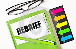 On a light background, multi-colored arrows, black glasses, a green notebook with a pen and a white card with the text DEBRIEF