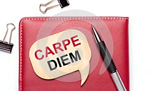 On a light background there are black paper clips, a pen, a burgundy notepad a wooden board with the text CARPE DIEM