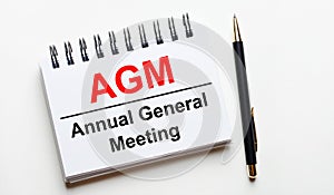 On a light background, a white notebook with are words AGM Annual General Meeting and a pen