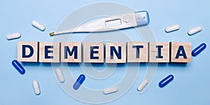 On a light blue background, an electronic thermometer, white and blue pills and wooden cubes with the inscription DEMENTIA. Medica