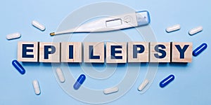 On a light blue background, an electronic thermometer, white and blue pills and wooden cubes with the inscription EPILEPSY.