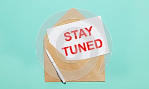 On a light blue background lies an open craft envelope, a white pen and a white sheet of paper with the text STAY TUNED