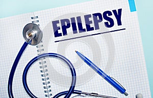 On a light blue background, an open notebook with the word EPILEPSY, a blue pen and a stethoscope. View from above. Medical