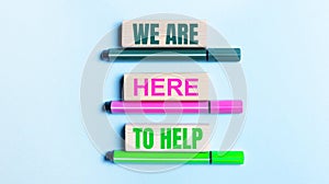 On a light blue background, there are three multi-colored felt-tip pens and wooden blocks with the WE ARE HERE TO HELP text