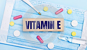 On a light blue disposable face mask there are tablets, a thermometer, an ampoule and a wooden block with the text VITAMIN E