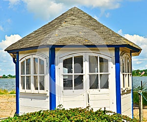 A light blue and white wooden small shed, pavilion, hexagonal at the waterside in summer