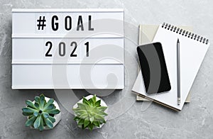 Light box with hashtag Goal 2021 near notebooks and modern smartphone on grey table, flat lay. New year targets