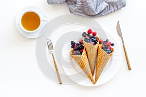 Light breakfast with fresh berries in waffle cones and cup of tea on served white table background top view