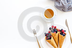 Light breakfast with fresh berries in waffle cones and cup of tea on served white table background top view mockup