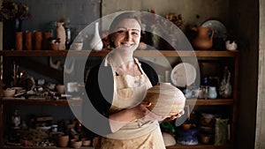 Light brown vase from clay in the female potter`s hands. Portrait of cheerful blonde woman holding a vase of clay. The