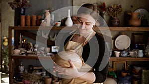 Light brown vase from clay in the female potter`s hands. A young and cheerful woman holding a vase of clay. The potter