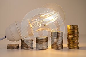 Light bulb, above stacks of coins. Electricity costs.