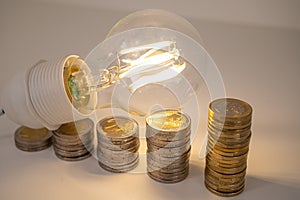 Light bulb, above stacks of coins. Electricity costs.