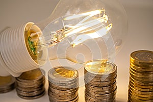 Light bulb, above stacks of coins. Electricity costs.