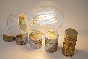 Light bulb, above stacks of coins. Electricity costs.