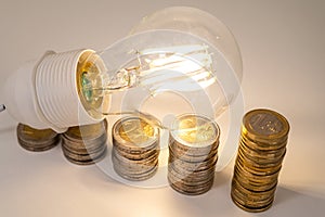 Light bulb, above stacks of coins. Electricity costs.