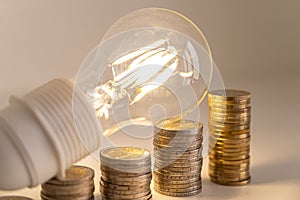 Light bulb, above stacks of coins. Electricity costs.