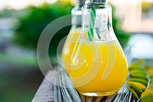 Light Bulb Glass Bottles with Fresh Orange Tropical Fruits Juice on Open Cafe Table at Sunset. Green Palm Trees Foliage Background