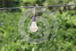 Light bulb hanging on the cord electric wire line cable with green nature tree bokeh lighting background