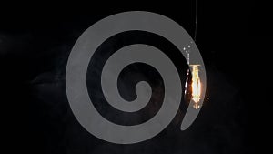 Light Bulb Hanging on Wire and Flickering on Black Background with Smoke
