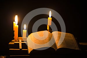 Light candle with holy bible and cross or crucifix on old wooden background in church.Candlelight and open book on vintage wood