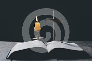 Light candle with holy bible and cross or crucifix on old wooden background in church.Candlelight and open book on vintage wood