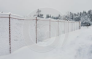 A light chain-link fence and a path in the village are covered with snow.