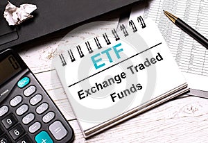 On a light-colored office table is a computer, a black calculator, a pen, and a notebook with the text ETF Exchange Traded Funds