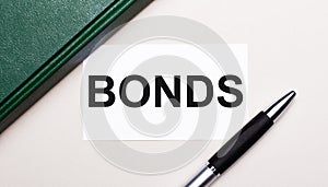 On a light gray background lies a pen, a green notebook and a white card with the text BONDS. Business concept