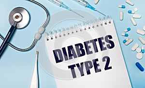 On a light gray background, a stethoscope, an electronic thermometer, pills, syringes and a notebook with the text DIABETES TYPE 2